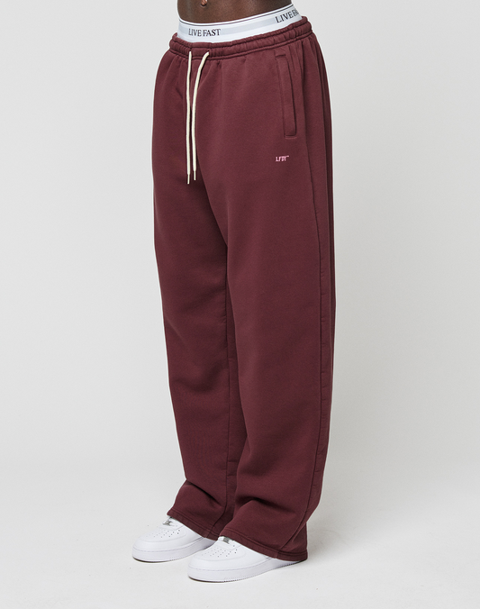 A person wears LIVE FAST DIE YOUNG Basic Wide Jogger in maroon with side pockets, white drawstrings, and a logo-print waistband, paired with white sneakers; only the lower body is visible against a plain background. | model