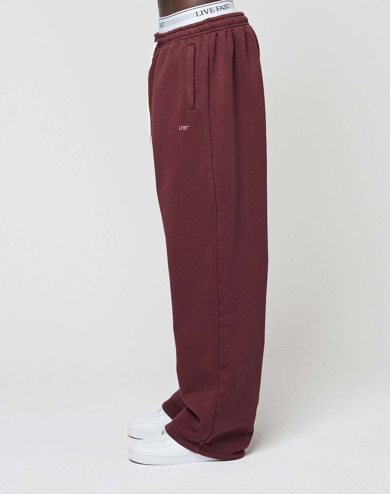 A person stands sideways in burgundy Basic Wide Jogger sweatpants by LIVE FAST DIE YOUNG, featuring seitliche Eingrifftaschen, a LIFT logo-print near the pocket, a white LIVE FAST waistband, and white sneakers against a plain background.