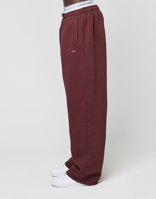 A person stands sideways in burgundy Basic Wide Jogger sweatpants by LIVE FAST DIE YOUNG, featuring seitliche Eingrifftaschen, a LIFT logo-print near the pocket, a white LIVE FAST waistband, and white sneakers against a plain background.