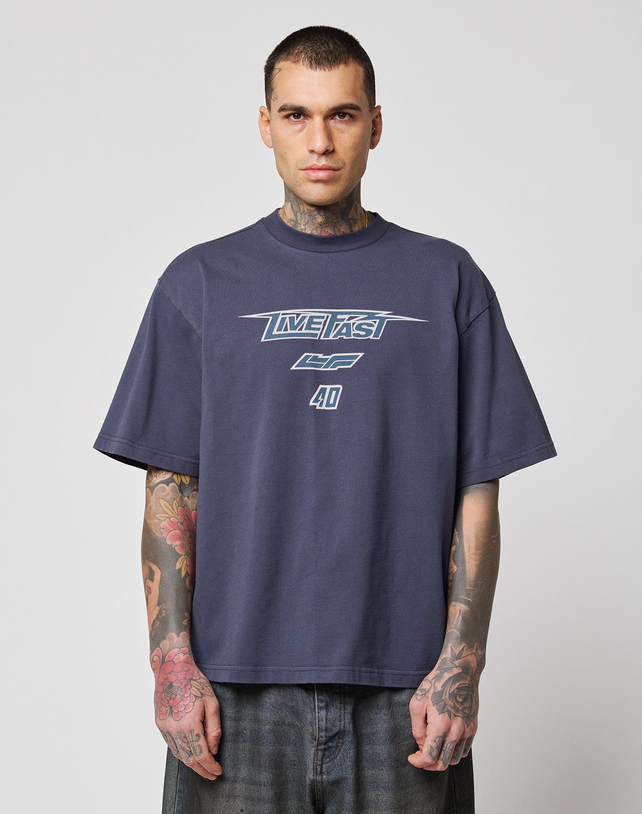 A person with short hair and visible tattoos wears an oversize fit, dark navy LFDY Triple Racing Tee by LIVE FAST DIE YOUNG with a Multilogo Frontprint reading LIVE FAST CF 40, standing against a plain light gray background. | model