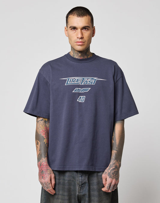 A person with short hair and visible tattoos wears an oversize fit, dark navy LFDY Triple Racing Tee by LIVE FAST DIE YOUNG with a Multilogo Frontprint reading LIVE FAST CF 40, standing against a plain light gray background.