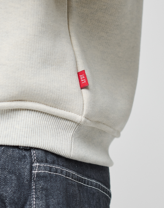 A close-up of a person wearing a light beige LIVE FAST DIE YOUNG LFDY Heavyweight Lounge Sweater, featuring an oversize fit and paired with dark denim jeans.