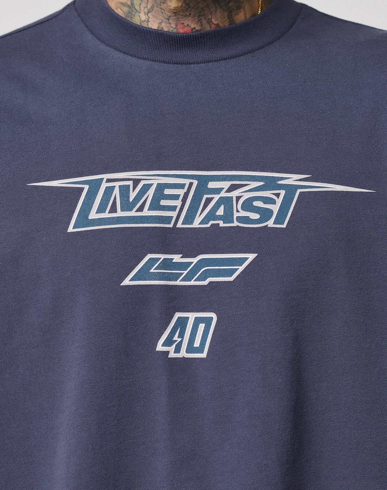 The LFDY Triple Racing Tee by LIVE FAST DIE YOUNG is a navy oversize t-shirt with “LIVE FAST” in stylized white and blue, LF and 40 below, plus multilogo frontprint. Made of 100% cotton. A tattooed neck appears at the image top.