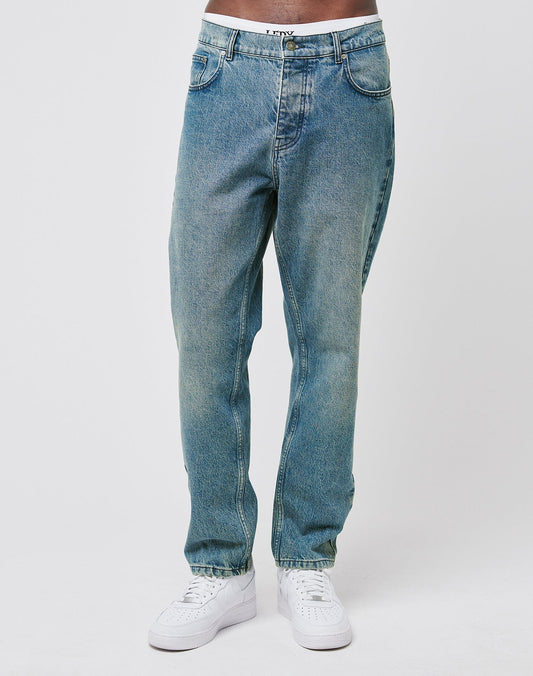 A model’s lower torso and legs are shown against a white background, wearing LIVE FAST DIE YOUNG Regular Denim jeans in light blue with a tapered fit and white sneakers.