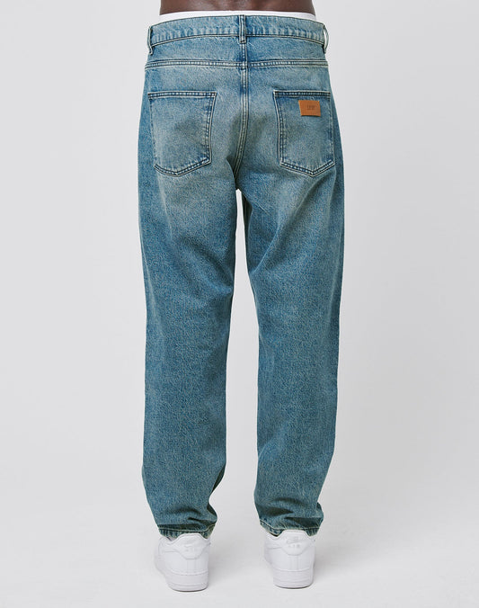 A person is seen from the back wearing LIVE FAST DIE YOUNG Regular Denim blue jeans with a tapered leg, two rear pockets, and a brown leather patch on the right pocket, paired with white sneakers against a white background.