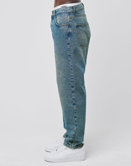 A person in LIVE FAST DIE YOUNG Regular Denim light blue, relaxed-fit jeans aus recycelter Baumwolle and white sneakers stands sideways against a plain white background.