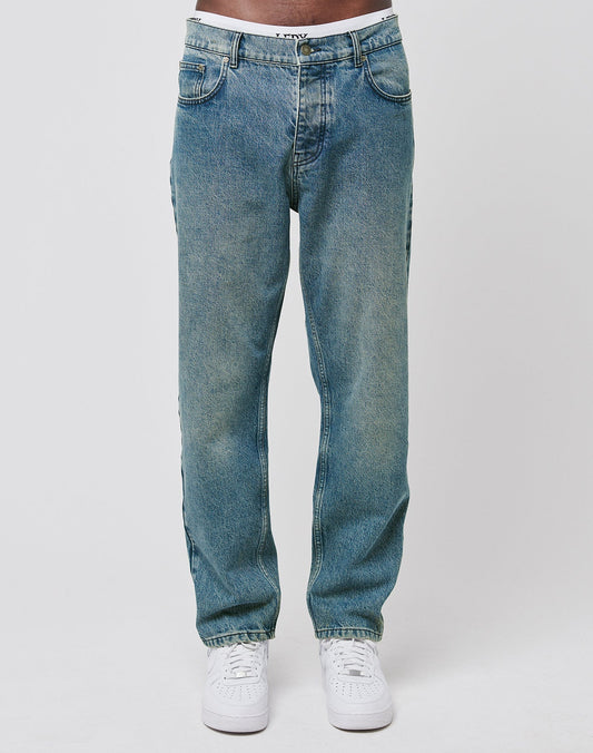 A person wears LIVE FAST DIE YOUNG Regular Denim jeans in light blue, featuring a regular fit and tapered leg made from recycelte Baumwolle, paired with white sneakers. The photo shows the jeans from the waist down against a plain white background.