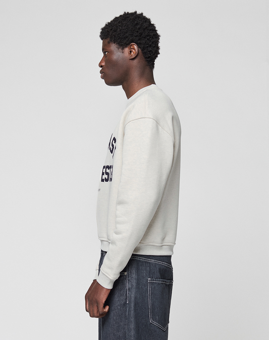 A person with short curly hair stands in profile, facing left, wearing the LIVE FAST DIE YOUNG LFDY Heavyweight Lounge Sweater in light gray and dark denim jeans against a plain light background.