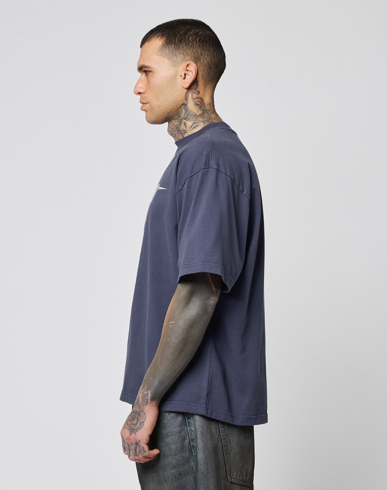 A person with short hair and tattoos stands in profile, wearing the LIVE FAST DIE YOUNG LFDY Triple Racing Tee—an oversize, dark blue 100% Baumwolle shirt with multilogo front print—plus blue jeans, against a plain light background.