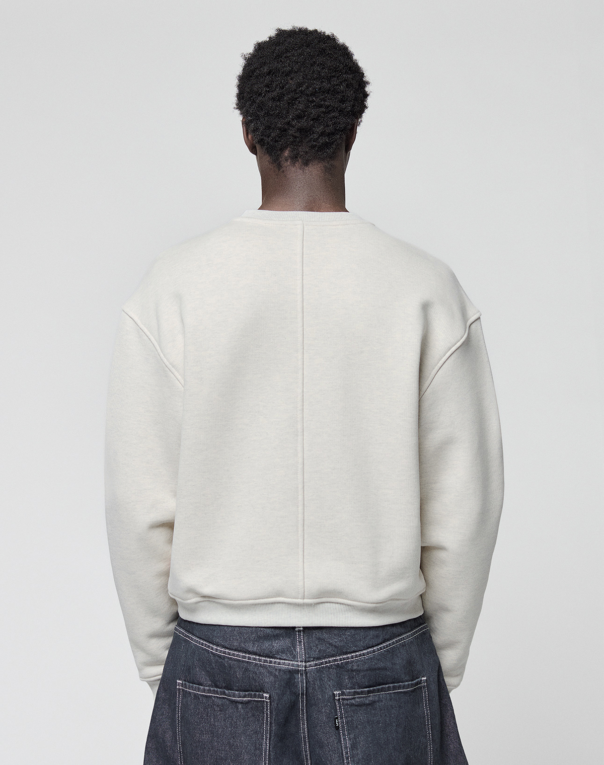 A person with short, curly hair stands with their back to the camera, wearing the LIVE FAST DIE YOUNG LFDY Heavyweight Lounge Sweater and dark denim jeans against a plain light gray background.