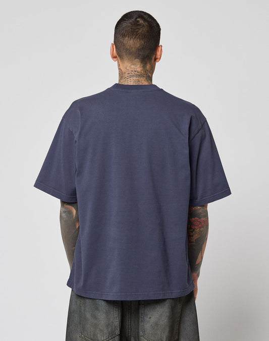 A person with short hair and tattoos stands with their back to the camera, wearing the LIVE FAST DIE YOUNG LFDY Triple Racing Tee in dark blue and dark pants against a plain background.
