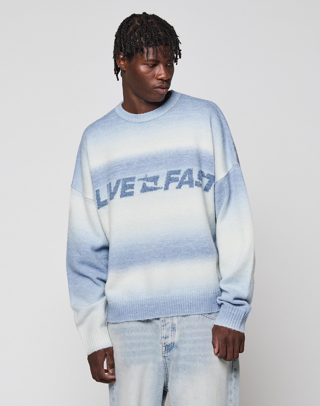 A person with short dreadlocks wears the LIVE FAST DIE YOUNG Live Fast Boucle Sweater, featuring an oversize blue and white gradient design and "LIVE FAST" text, paired with light-wash jeans against a plain light background.