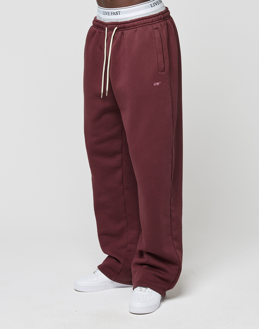 A person wears the LIVE FAST DIE YOUNG Basic Wide Jogger in maroon with side pockets, drawstrings, a white LIVE FAST waistband, and white sneakers, standing against a plain white background.