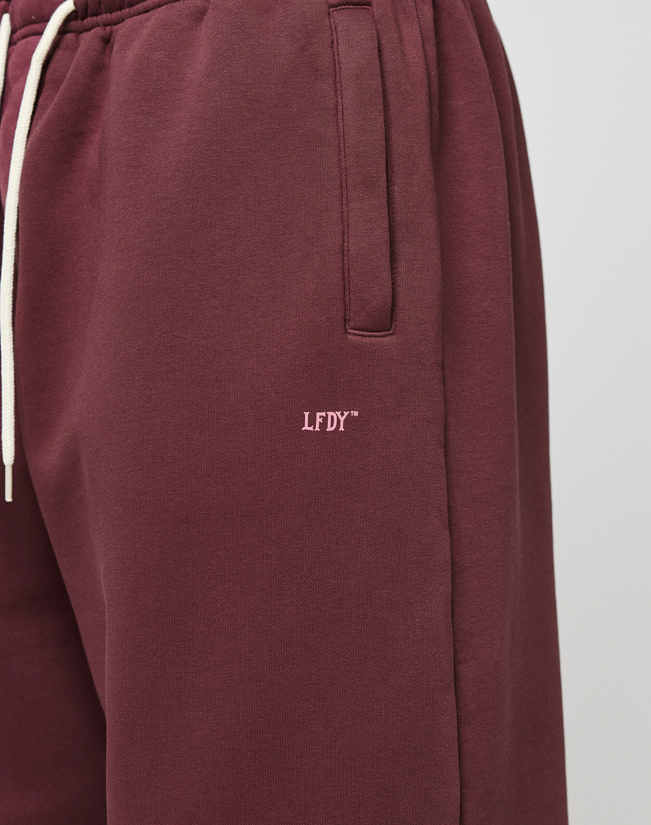 Close-up of a person wearing the LIVE FAST DIE YOUNG Basic Wide Jogger in maroon with wide fit, side pockets, white drawstrings, and small white LFDY™ logo embroidered on the left thigh.