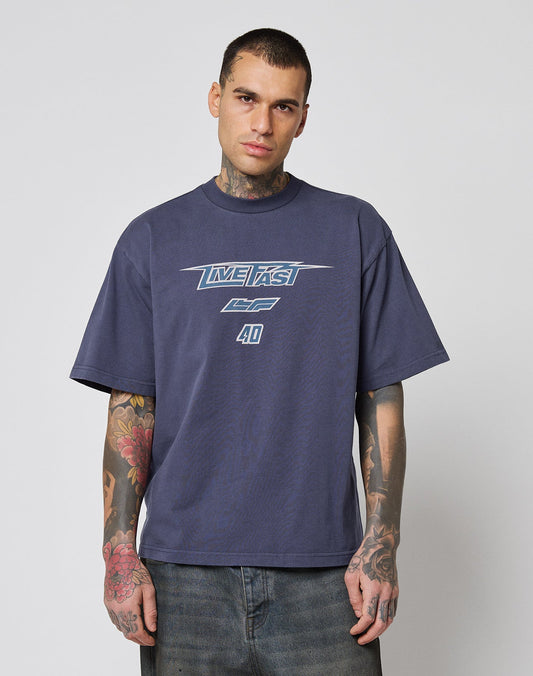 A tattooed man stands against a plain background, wearing the LFDY Triple Racing Tee from LIVE FAST DIE YOUNG—an oversize fit, dark blue 100% cotton shirt with multilogo front print featuring the text LIVE FAST 4D.