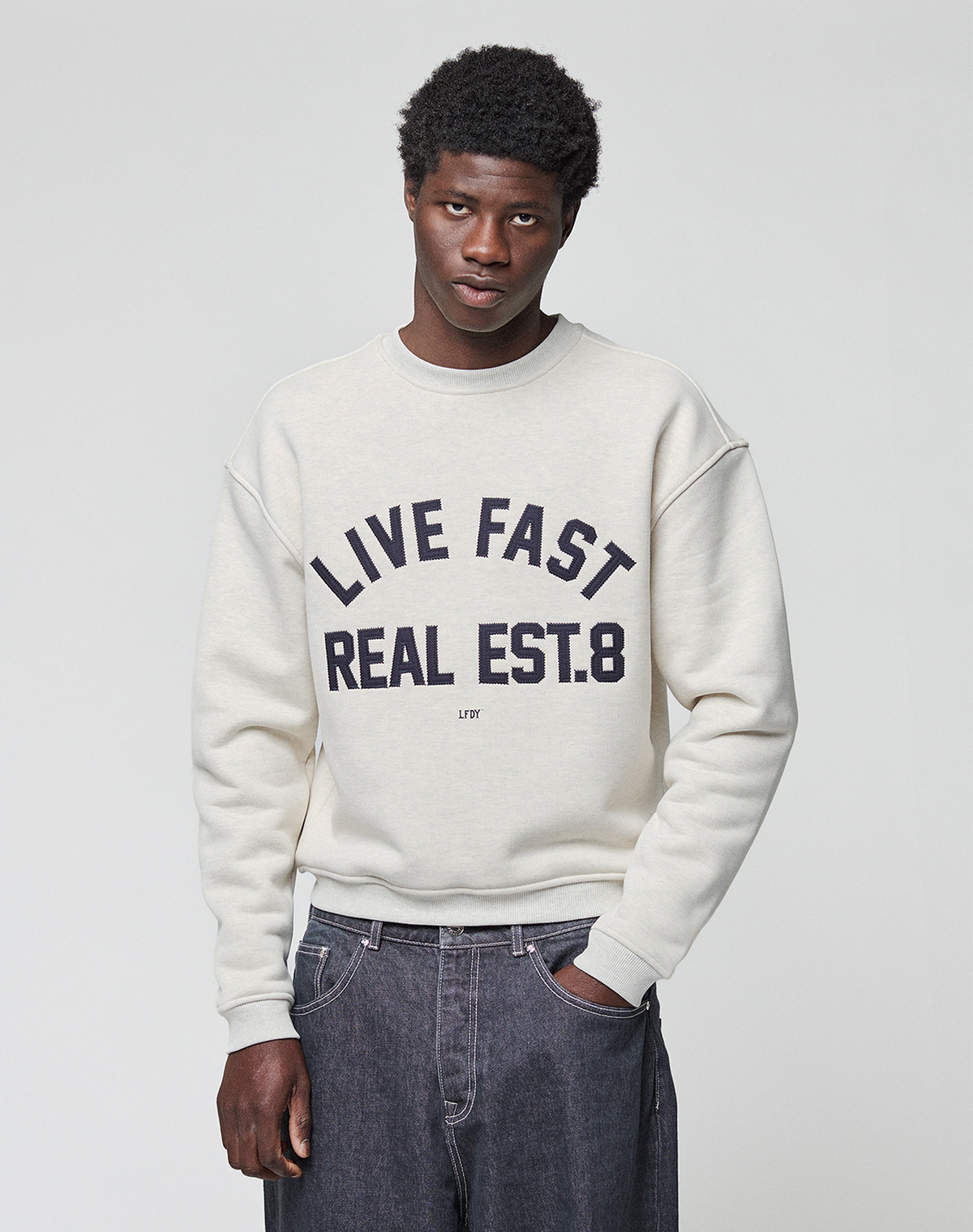 A person with short, curly hair and a neutral expression wears a LIVE FAST DIE YOUNG LFDY Heavyweight Lounge Sweater with "LIVE FAST REAL EST.8" in bold over dark jeans, standing against a plain background. The sweater is made from 100% Baumwolle.