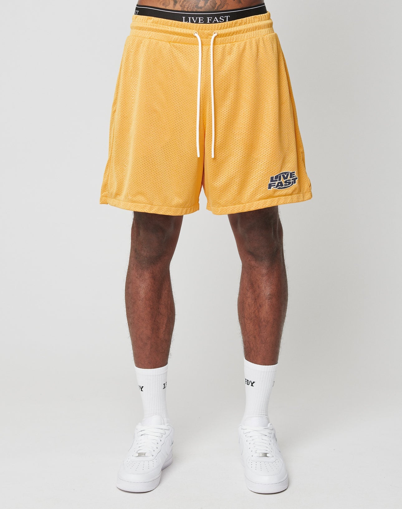 A person, shown from the waist down, wears LFDY Oval Mesh Shorts by LIVE FAST DIE YOUNG with an elastic waistband and LIVE FAST text, paired with white socks and sneakers, standing against a plain light background.