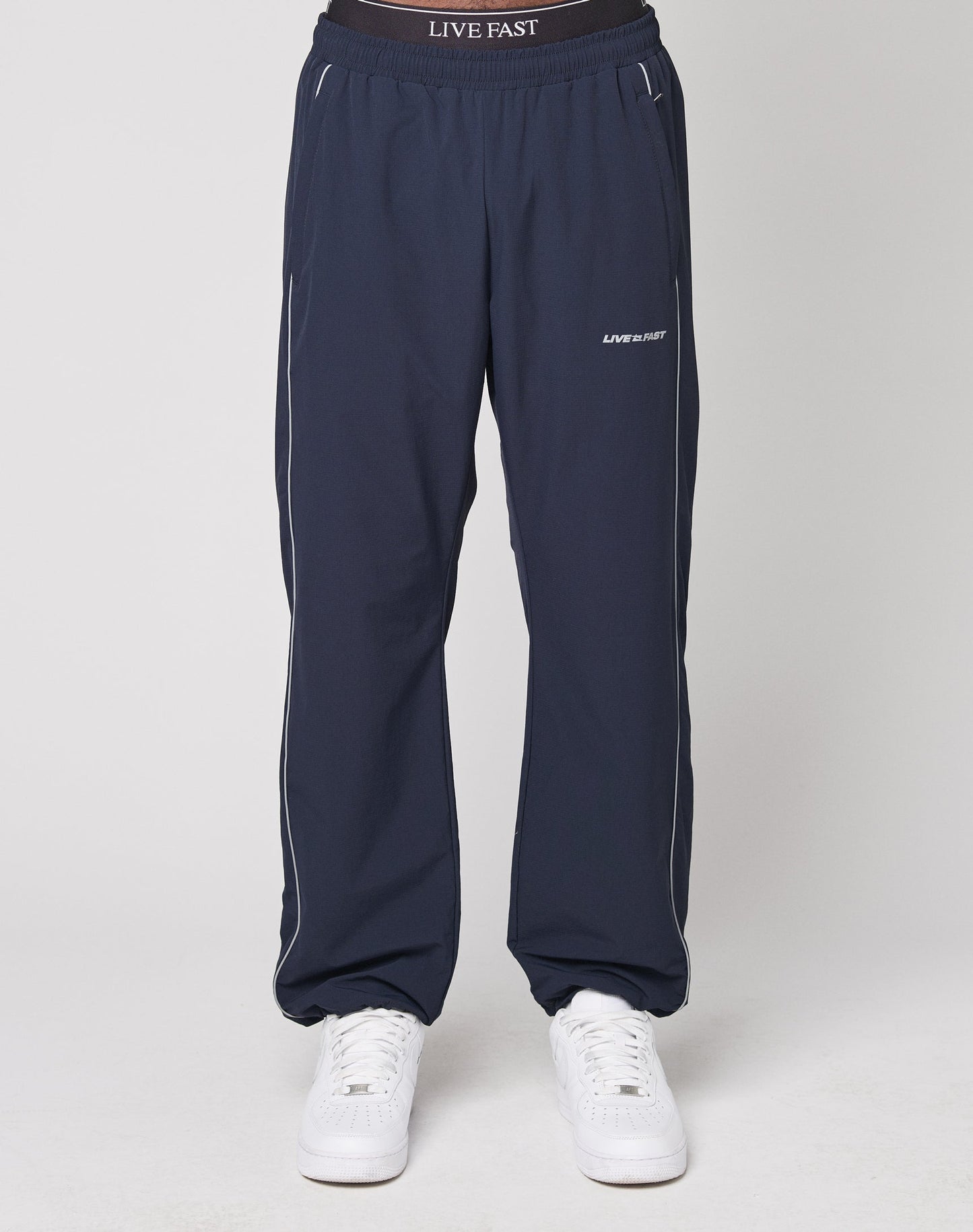 Someone stands against a plain background wearing LIVE FAST DIE YOUNG’s navy blue Live Fast Trackpants, featuring a reflektierender Logo Print on the waistband and an elastikbund for comfort, paired with white sneakers.