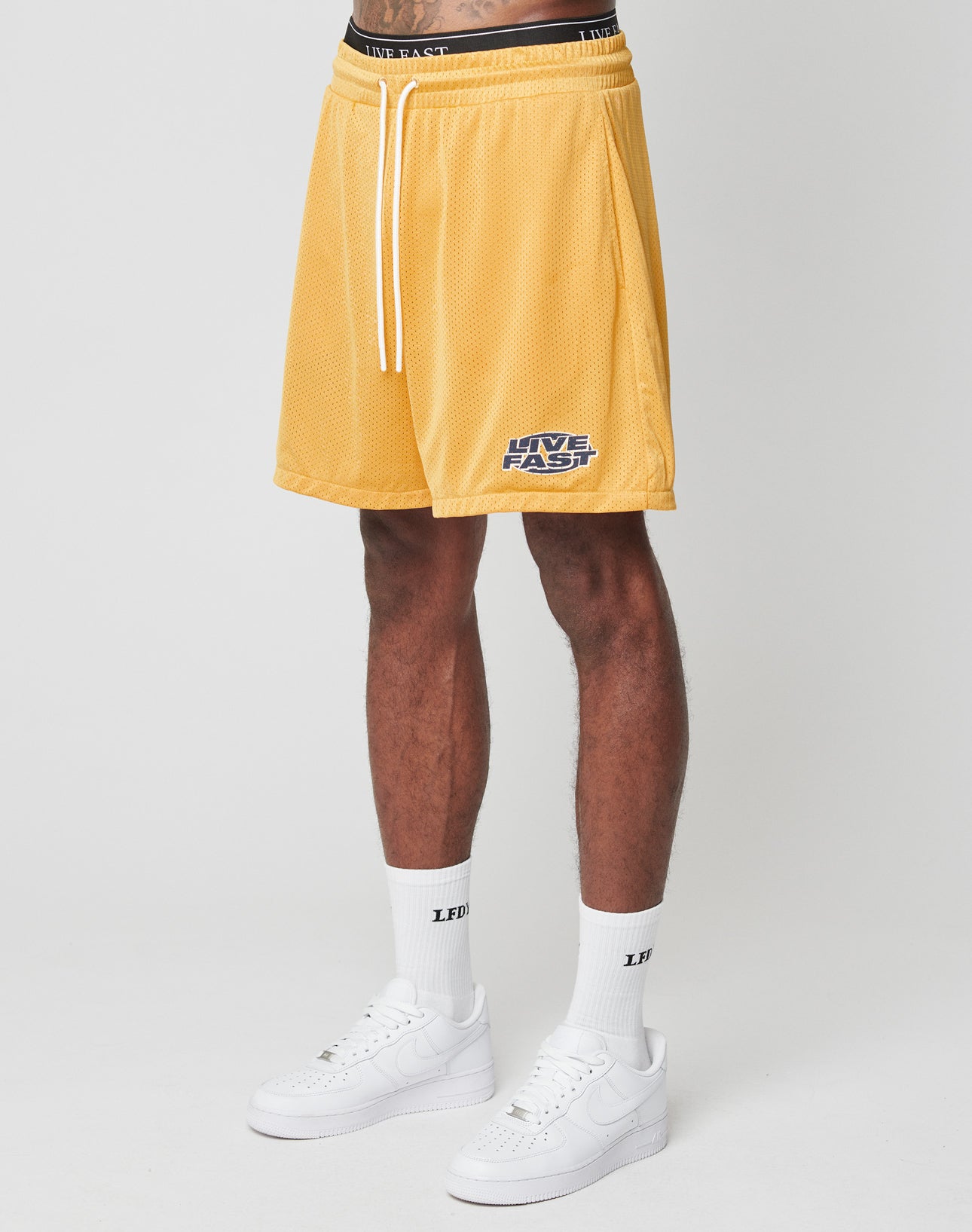 A person wears yellow LFDY Oval Mesh Shorts by LIVE FAST DIE YOUNG, featuring an elastic waistband and LIVE FAST text, paired with white socks with black LFDY text and white sneakers, standing against a plain white background. | model