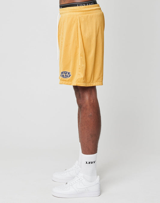 A person wears LIVE FAST DIE YOUNG LFDY Oval Mesh Shorts in yellow with an elastic waistband, white LFDY socks, and white sneakers, standing sideways against a plain white background. The polyester shorts have LIVE FAST text near the hem.