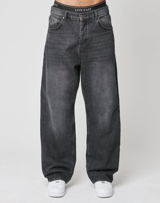 A person in LFDY Baggy Denim by LIVE FAST DIE YOUNG, dark grey and 100% cotton, paired with white sneakers stands facing forward. The black LIVE FAST waistband is visible above the jeans against a plain white background.
