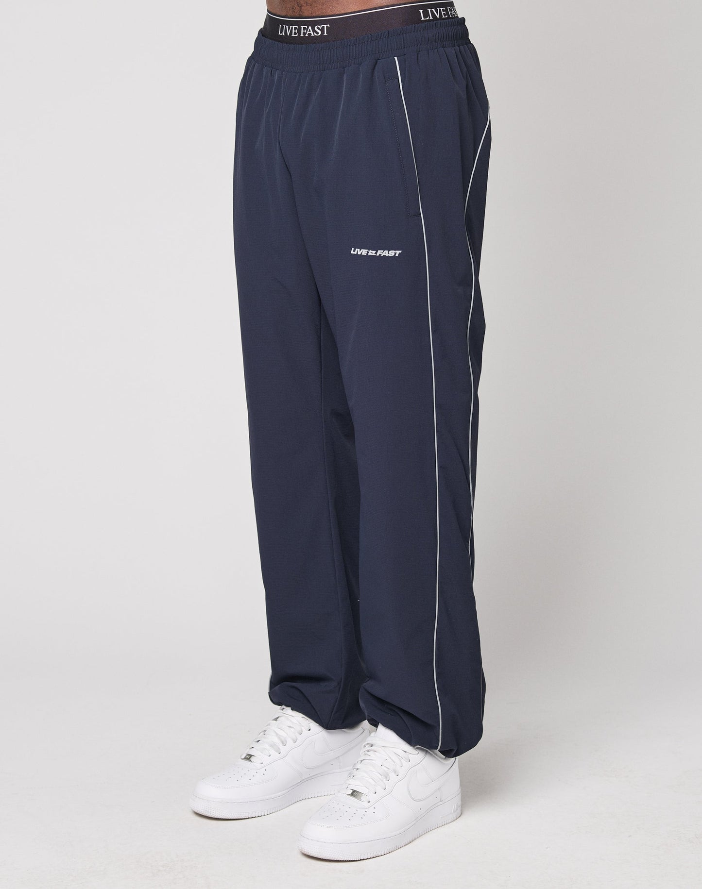 A person wearing LIVE FAST DIE YOUNG Live Fast Trackpants in navy blue wide fit with white piping, elasticated waistband, reflective logo prints on waist and thigh, and white sneakers, stands against a plain light background.