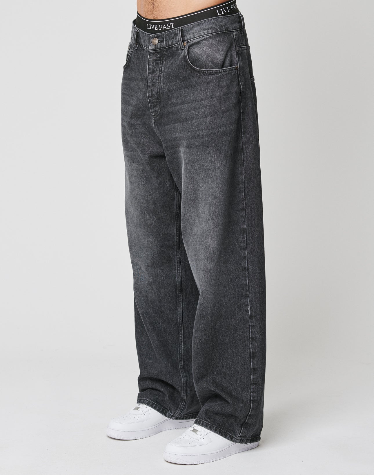 A person wearing LFDY Baggy Denim by LIVE FAST DIE YOUNG—baggy, faded black jeans with a wide straight leg made from 100% cotton—and white sneakers stands against a plain white background. The waistband reads LIVE FAST. | model