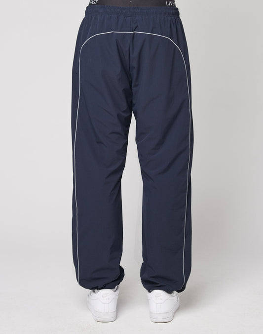 A person is seen from behind wearing LIVE FAST DIE YOUNGs Live Fast Trackpants in navy with white piping, a reflective logo print, and an elastic waistband in a wide fit, paired with white sneakers against a plain white background.