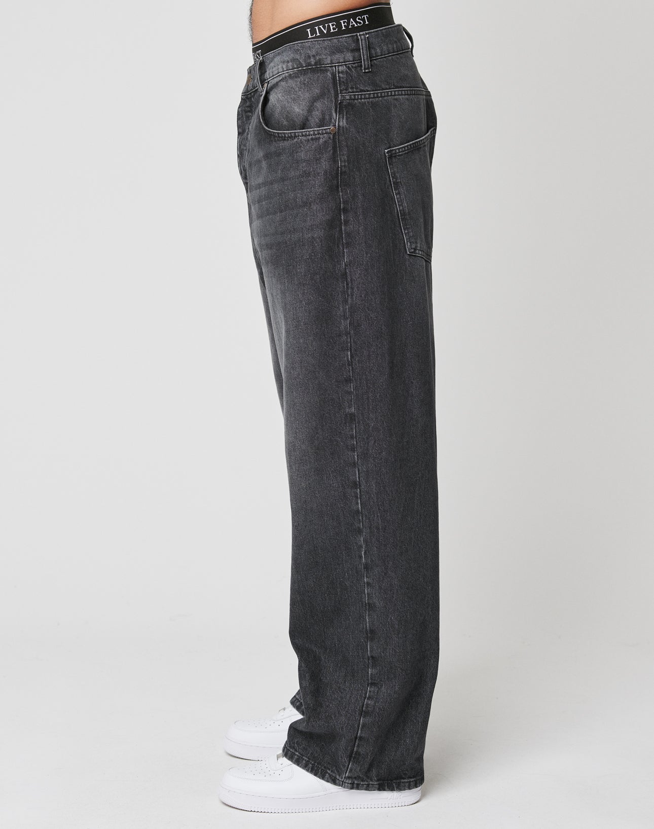 A person stands sideways in LFDY Baggy Denim by LIVE FAST DIE YOUNG—loose black jeans with a straight leg and visible LIVE FAST waistband—paired with white sneakers, against a plain white background.