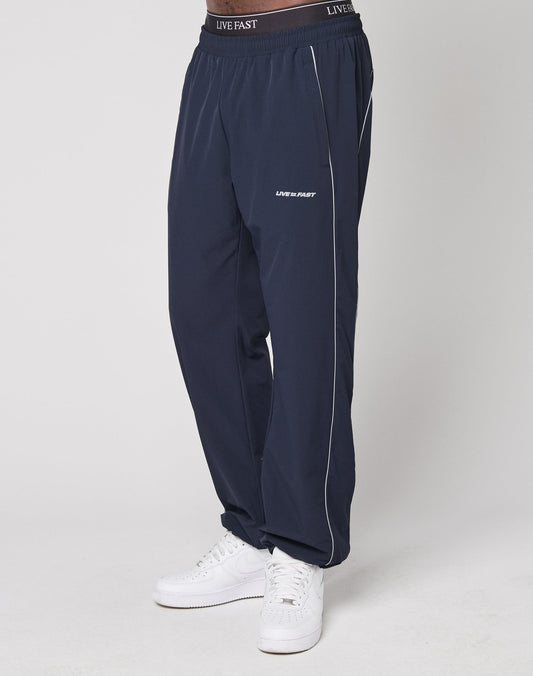 Wearing LIVE FAST DIE YOUNG Live Fast Trackpants in navy blue with white piping, wide fit, black elastic waistband featuring LIVE FAST, and reflective logo print on the thigh, paired with white sneakers against a plain light background.