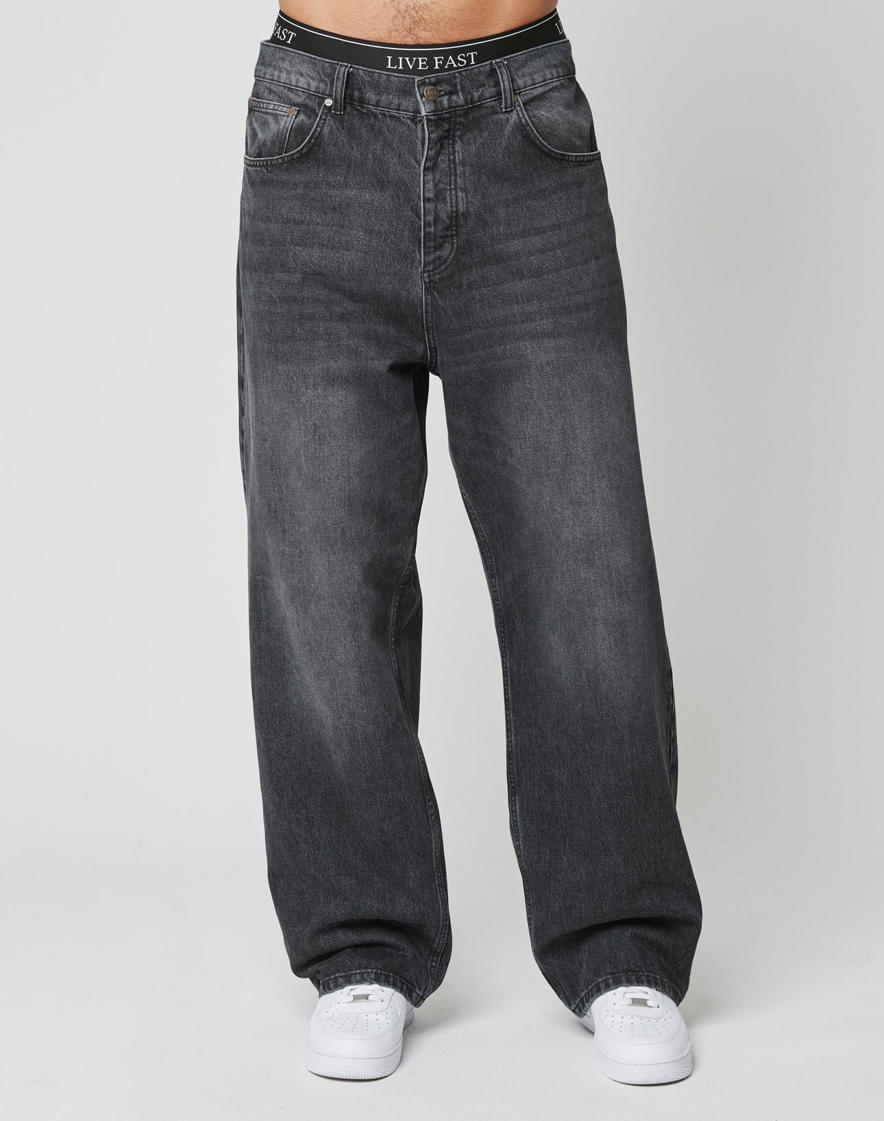 A person wears LFDY Baggy Denim by LIVE FAST DIE YOUNG—baggy fit, dark gray jeans with a wide straight leg and LIVE FAST on the 100% Baumwolle waistband—paired with white sneakers against a plain light background.