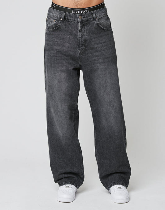 A person wears LFDY Baggy Denim by LIVE FAST DIE YOUNG—baggy fit, dark gray jeans with a wide straight leg and LIVE FAST on the 100% Baumwolle waistband—paired with white sneakers against a plain light background.