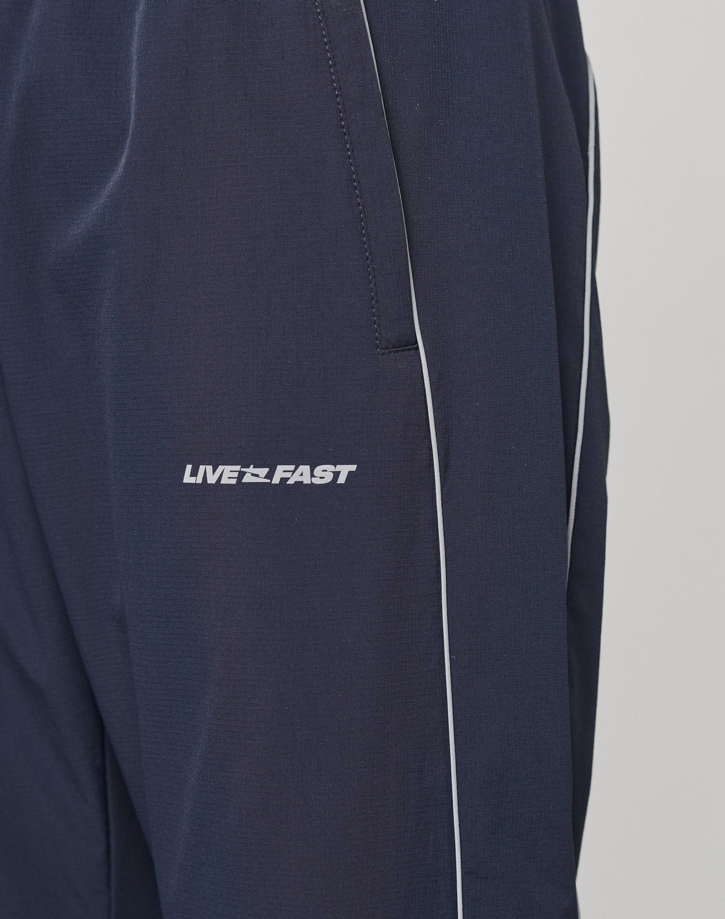 Close-up of LIVE FAST DIE YOUNGs Live Fast Trackpants in navy blue with white piping, a reflective LIVE FAST logo print on the left leg, and an elastic waistband for extra comfort.