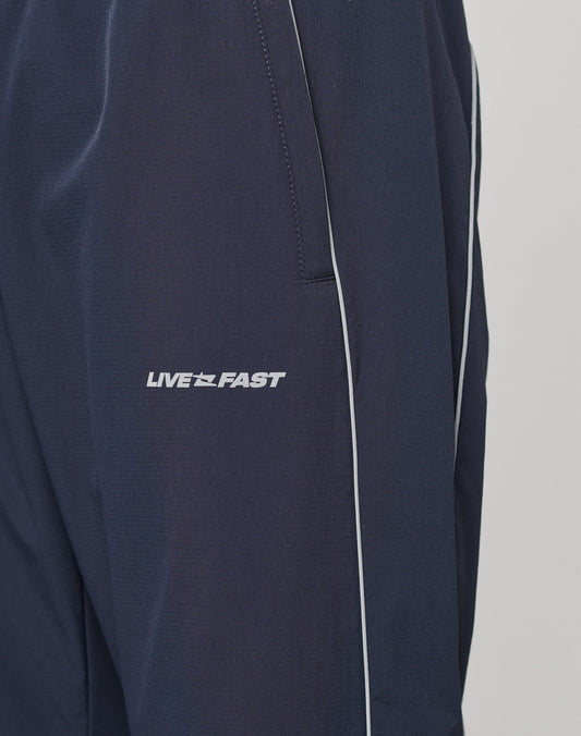 Close-up of LIVE FAST DIE YOUNGs Live Fast Trackpants in navy blue with white piping, a reflective LIVE FAST logo print on the left leg, and an elastic waistband for extra comfort.