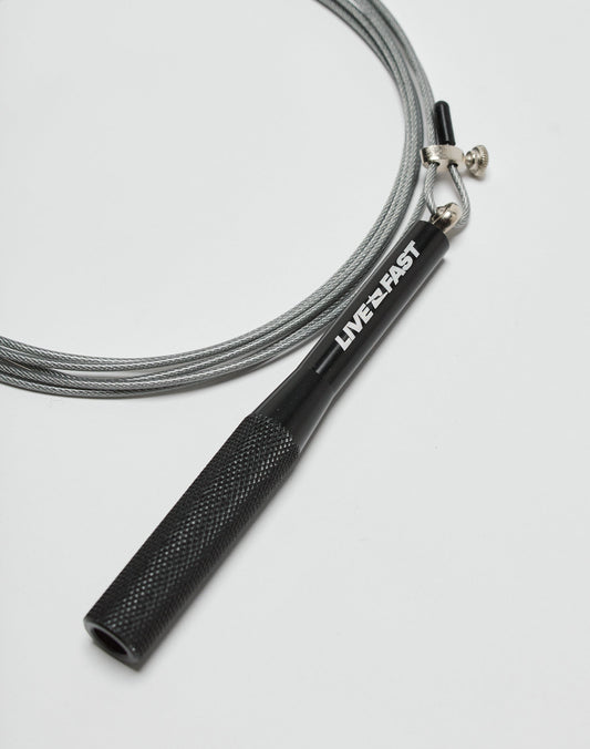 The Live Fast Performance Jump Rope by LIVE FAST DIE YOUNG features a black handle with LIVE•FAST in white and an adjustable wire cable, shown on a white surface.