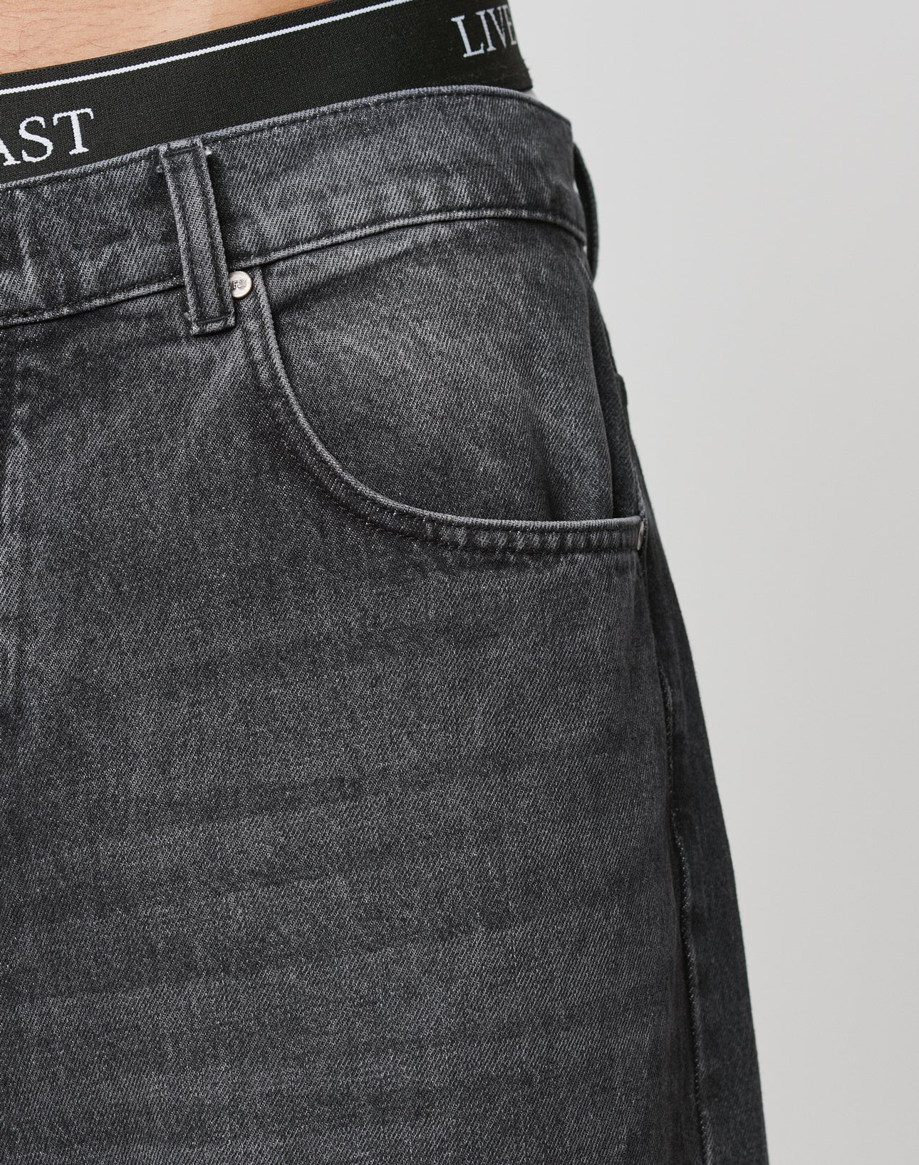 Close-up of a person wearing LFDY Baggy Denim by LIVE FAST DIE YOUNG, showcasing the faded black jeans baggy fit with focus on the front pocket and waistband text, set against a plain light background.
