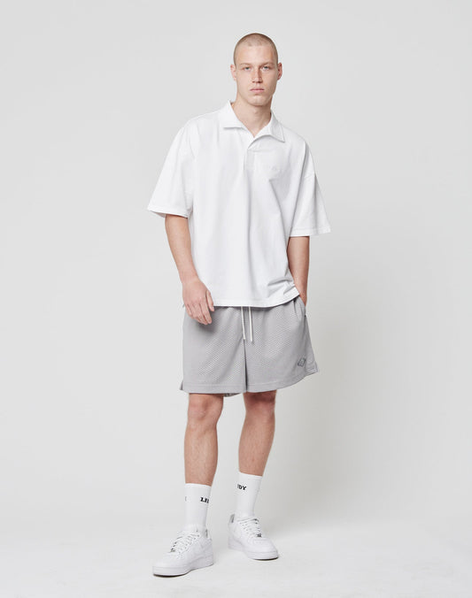 A young man with short hair and a neutral expression stands against a white background, wearing LIVE FAST DIE YOUNG Basic Mesh Shorts in light gray polyester, a loose white polo with logo patch, white crew socks, and sneakers.