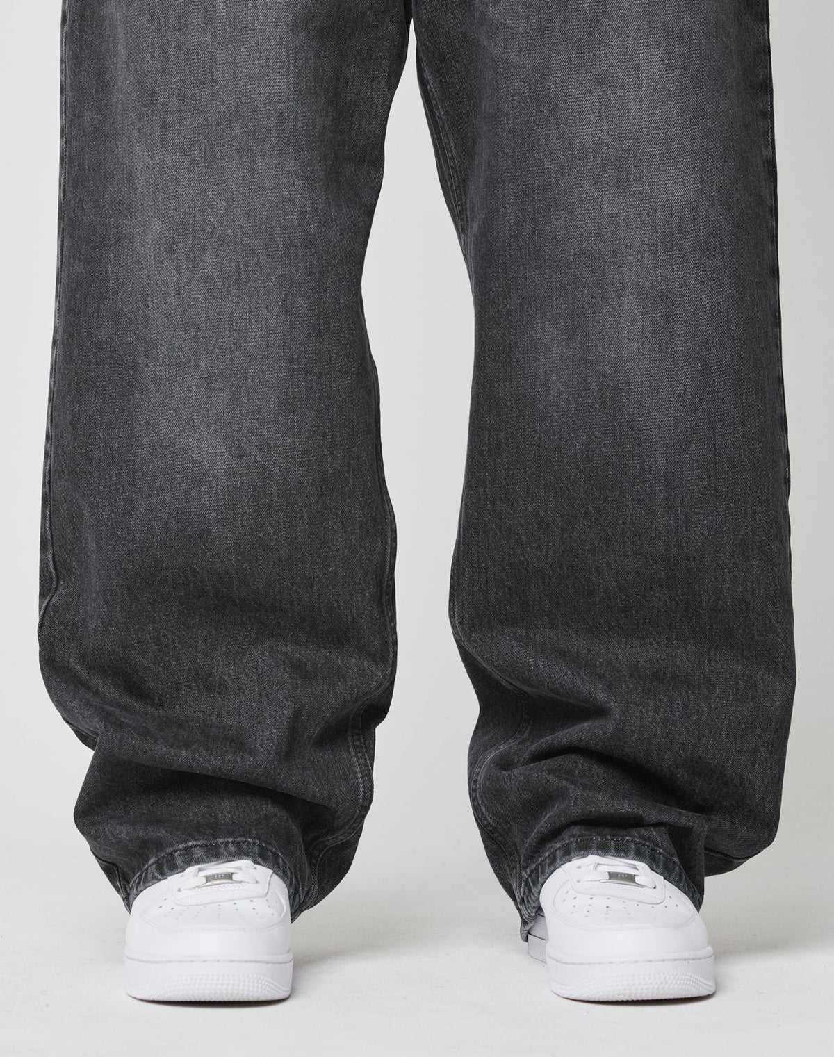 A person stands on a white surface, wearing LIVE FAST DIE YOUNG’s LFDY Baggy Denim in dark gray and white sneakers. The photo is cropped to display only the legs and feet.