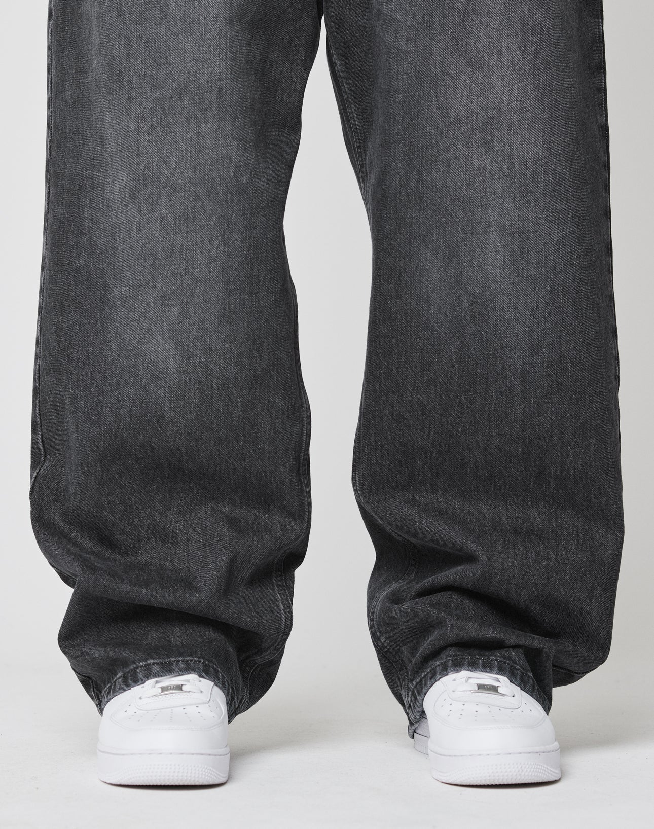 A person stands on a white surface, wearing LIVE FAST DIE YOUNG’s LFDY Baggy Denim in dark gray and white sneakers. The photo is cropped to display only the legs and feet.