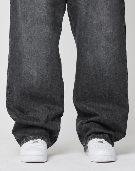 A person stands on a white surface, wearing LIVE FAST DIE YOUNG’s LFDY Baggy Denim in dark gray and white sneakers. The photo is cropped to display only the legs and feet.