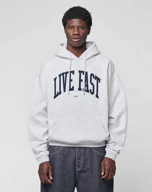 A young man faces the camera in a light gray Live Fast College Hooded sweatshirt by LIVE FAST DIE YOUNG, featuring a bold "LIVE FAST" front print, paired with dark jeans against a plain light gray background. | model