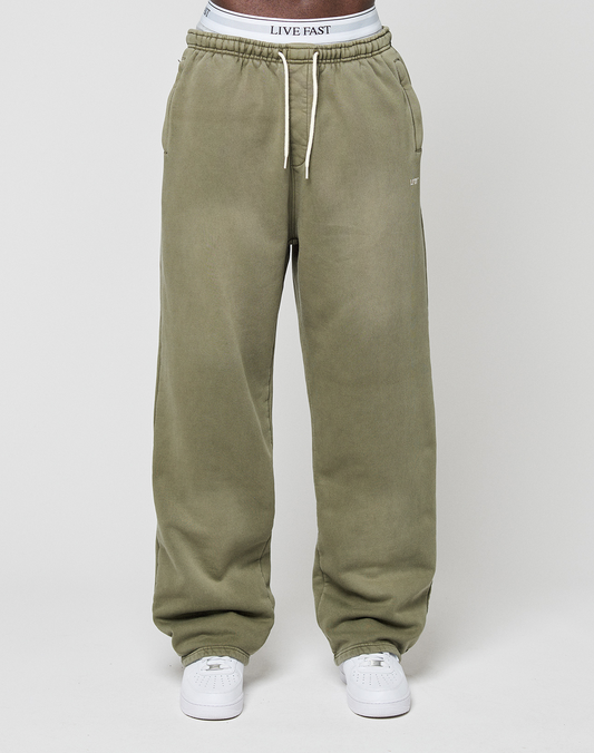 Someone wearing LIVE FAST DIE YOUNG Basic Wide Jogger sweatpants in olive green with drawstrings, paired with white sneakers and gray LIVE FAST DIE YOUNG underwear waistband visible above the pants.