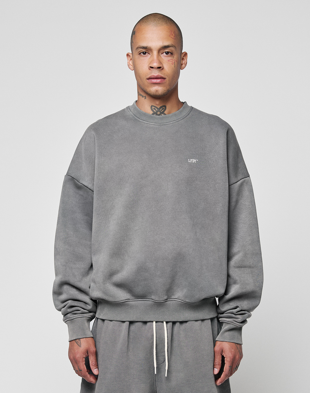 A person with a shaved head and tattoos wears the LIVE FAST DIE YOUNG Basic Chest Sweater—an oversize fit, faded gray sweatshirt made from soft Baumwolle—along with matching sweatpants, against a plain light gray background. | model