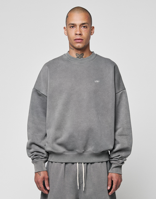 A person with a shaved head and tattoos wears the LIVE FAST DIE YOUNG Basic Chest Sweater—an oversize fit, faded gray sweatshirt made from soft Baumwolle—along with matching sweatpants, against a plain light gray background. | model