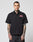 A man with tattoos models the LFDY Workwear Shirt by LIVE FAST DIE YOUNG, a black oversized cotton short-sleeve button-up with two chest pockets and a red patch on the left, standing facing forward against a plain light background.