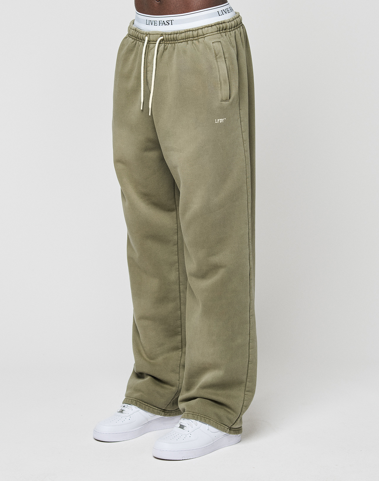 A person wears the LIVE FAST DIE YOUNG Basic Wide Jogger—olive green, wide fit, with “LIVE FAST” on the waistband and left pocket—paired with white sneakers, standing against a plain white background. | model