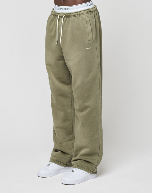 A person wears the LIVE FAST DIE YOUNG Basic Wide Jogger—olive green, wide fit, with “LIVE FAST” on the waistband and left pocket—paired with white sneakers, standing against a plain white background. | model