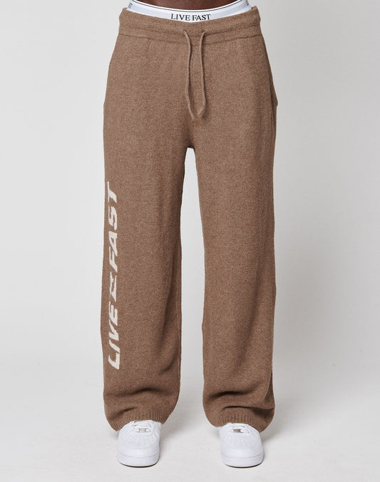 A person wearing LIVE FAST DIE YOUNG's brown Live Fast Boucle Pants with "LIVE FAST" in white on one straight leg, white sneakers, and a visible elastic waistband that also says "LIVE FAST.