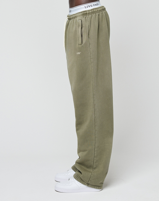A person in LIVE FAST DIE YOUNG’s Basic Wide Jogger (olive green) with a white elastic waistband and white sneakers stands sideways against a plain white background.