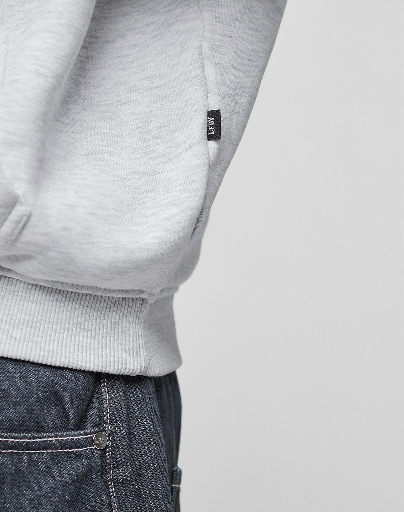 Close-up of a person wearing the LIVE FAST DIE YOUNG Live Fast College Hooded in light gray paired with blue jeans. Only the lower part of the hoodie and the jeans' waistband are visible.